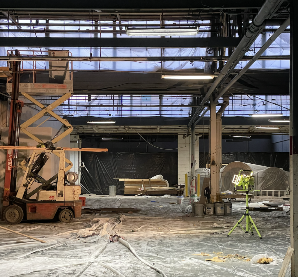 Natural light coming through banks of windows in raised industrial ceilings. Scissor lift on the left. Poly on the floors.