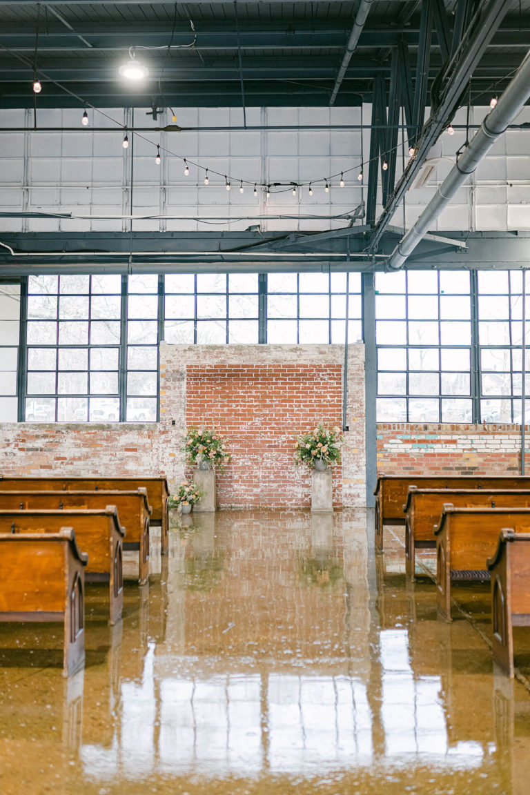 Ceremony Aisle with wooden pews on each side and brick wall as back drop. Beautiful wedding floral arrangements up front.