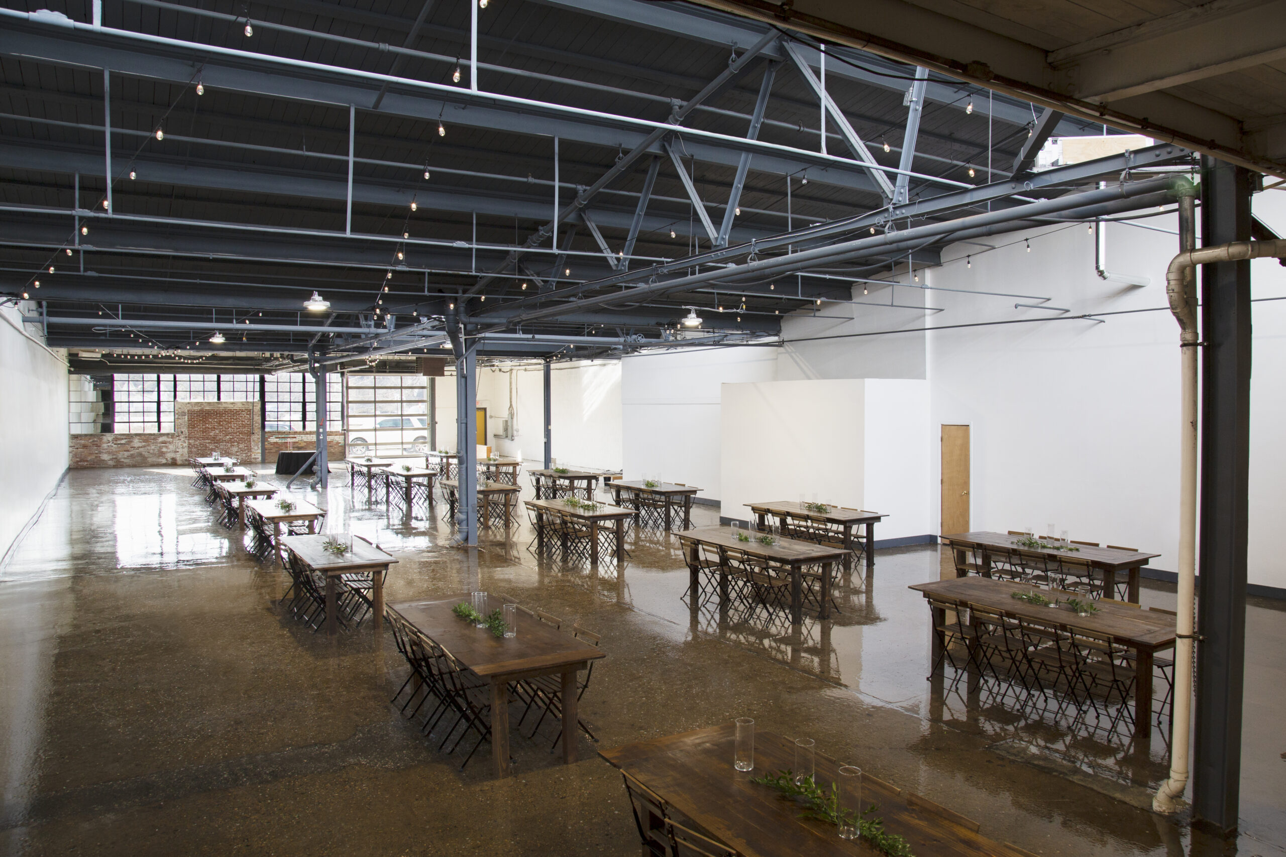 View of an 8000 square foot event venue space with high ceilings, concrete floor. Set up with farm tables and wooden chairs.