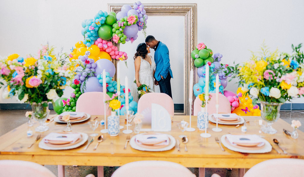 Light wood farm table with pink arm chairs surrounding. Bright table scape with taper candles and place settings. Giant frame in the background with colorful balloons and a bride and groom