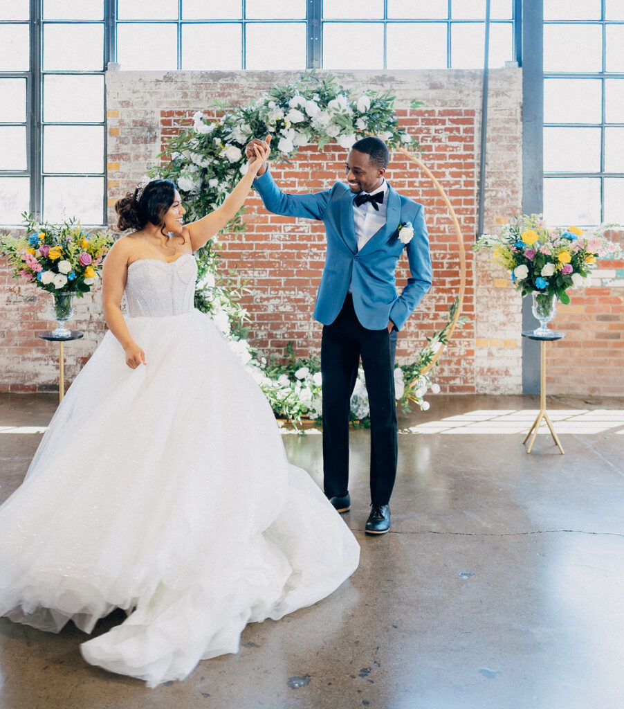 Bride in a white dress bring shown off by groom in a blue suite. Circle backdrop with florals against an exposed brick wall and industrial windows.