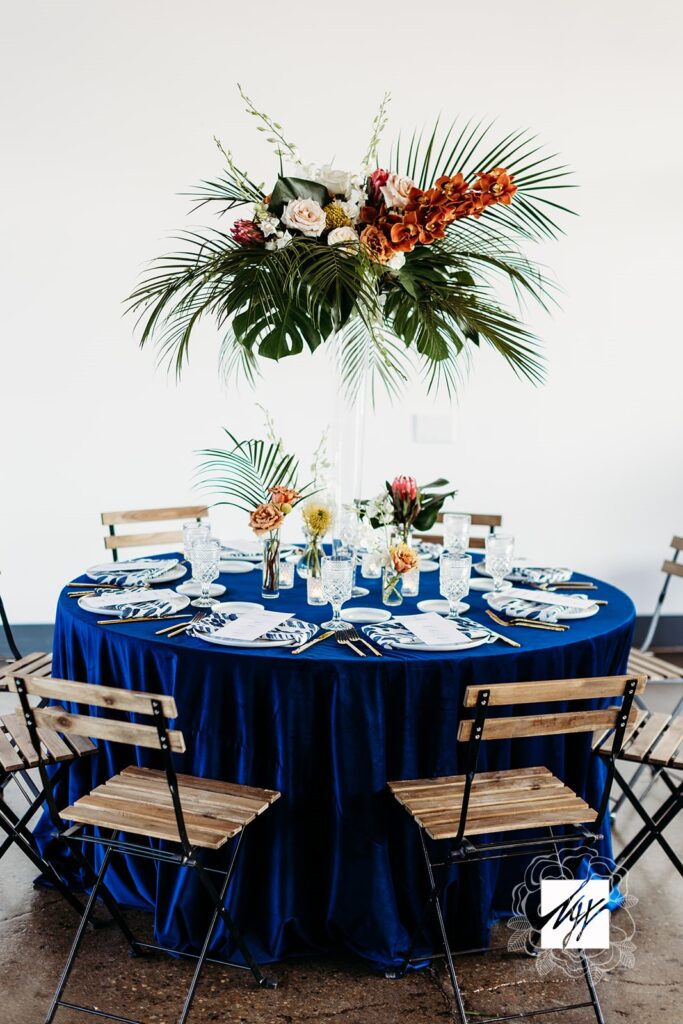 Round table with blue linen, white china, brown wooden chairs with black metal, and tropical florals