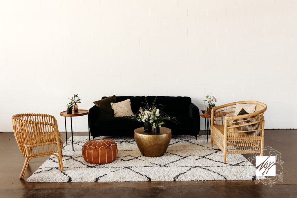 Soft seating area with a black sofa, black and white shaggy rug, two rattan chairs and gold accent tables. Against a white wall