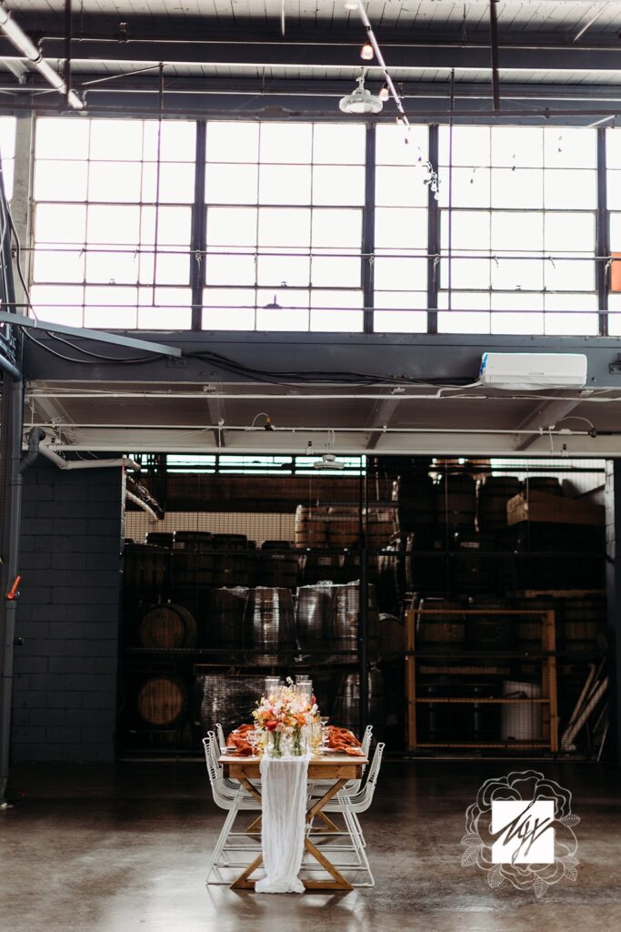 Dinner party set up with a farm table, white wire chairs, tablescape with orange hues on a concrete floor with industrial windows at the top