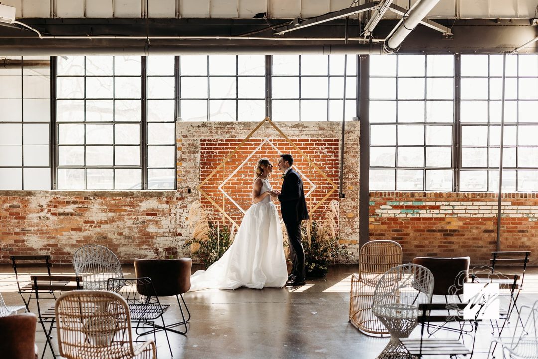 Wedding Ceremony with bride and groom against an exposed brick wall and banks of industrial windows. Concrete floor with mismatched chairs